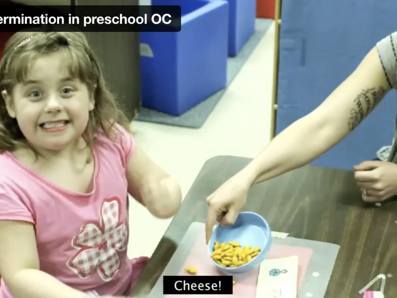 SD in Preschool Screen grab from the Self-determination in Preschool video showing a young female student smiling at the camera while her teacher, sitting close by, shares some goldfish crackers with her.