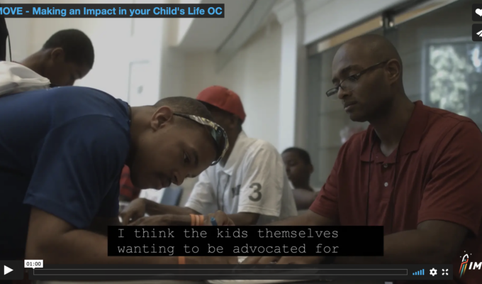 Screen capture of video screen showing two men at a table. One is filling out a form and the other is assisting.