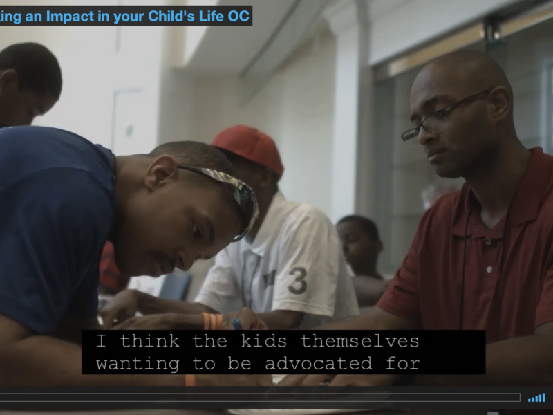 Screen capture of video screen showing two men at a table. One is filling out a form and the other is assisting.