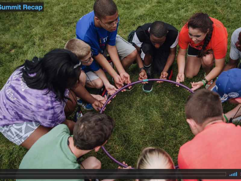 Screen capture from the 2016 Summit Recap video featuring a group of youth squatting in a circle and holding a hula-hoop in their hands.