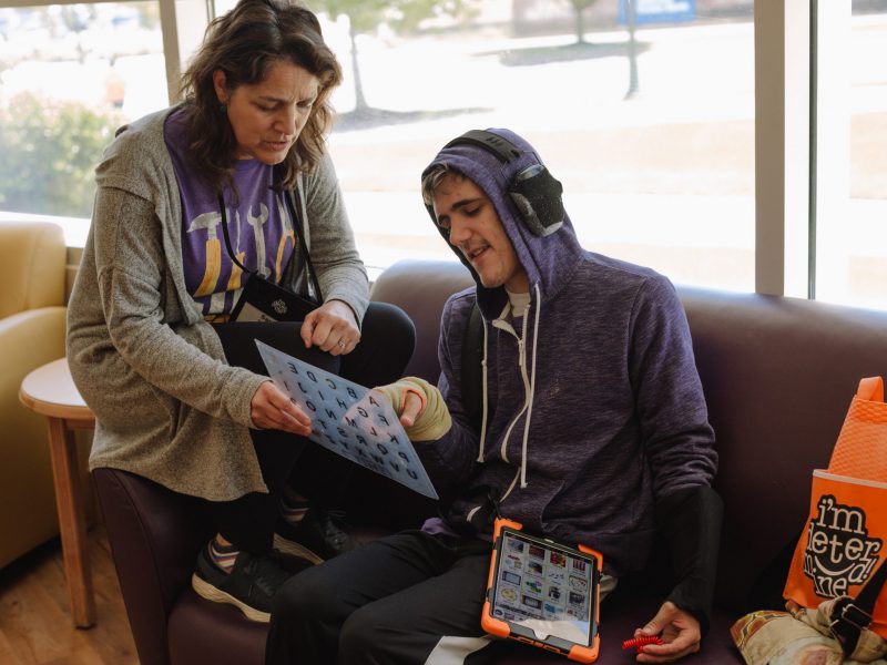 a teacher helps a teenage boy communicate with a letter-board