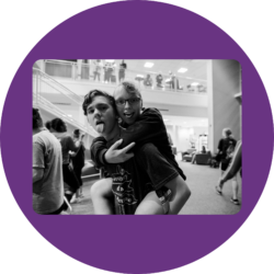 Two participants during the social at the Youth & Family Summit. One is giving the other a piggy-back ride and they are both smiling with tongues out.